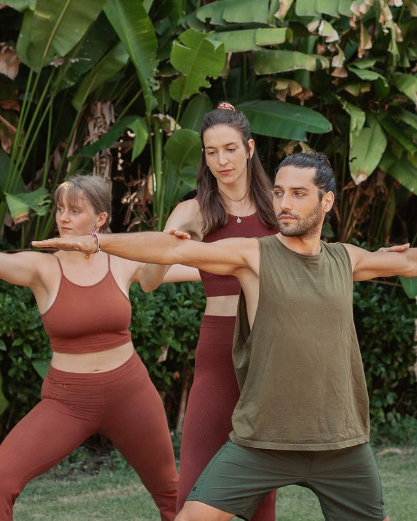 A yoga teacher leads two students through warrior two on the grass in a tropical garden.