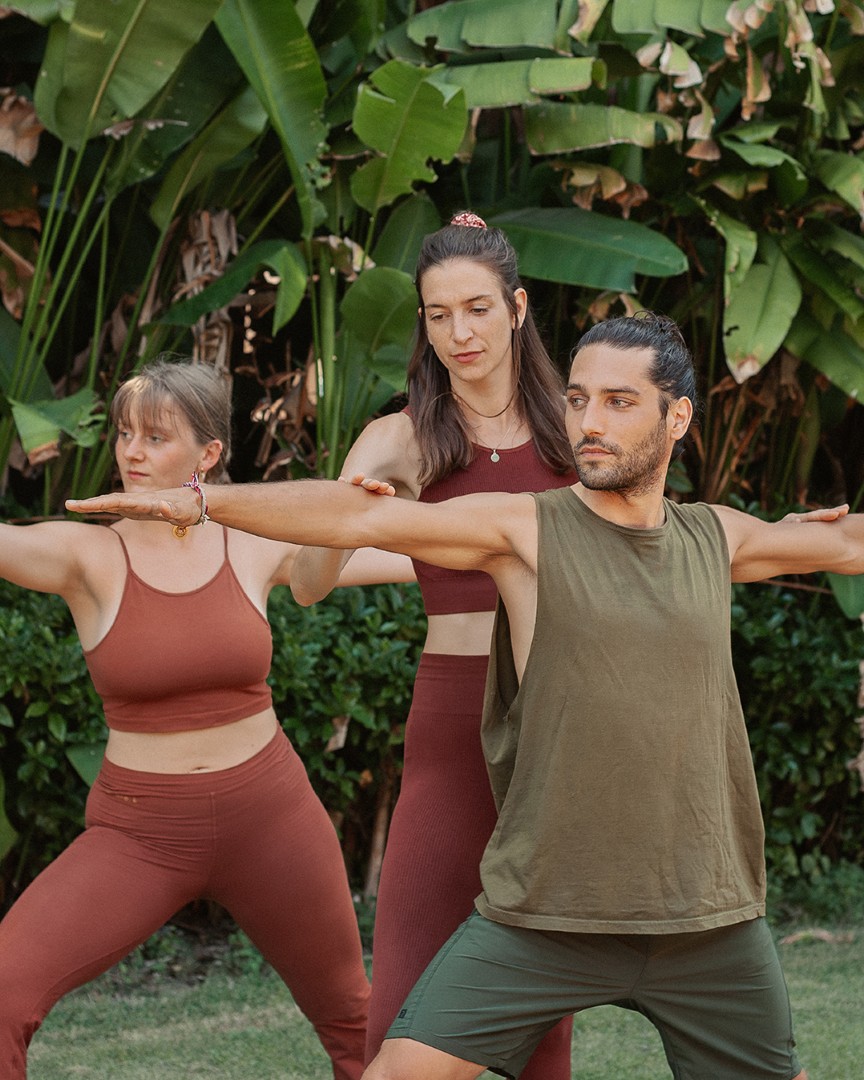 A yoga teacher leads two students through warrior two on the grass in a tropical garden.