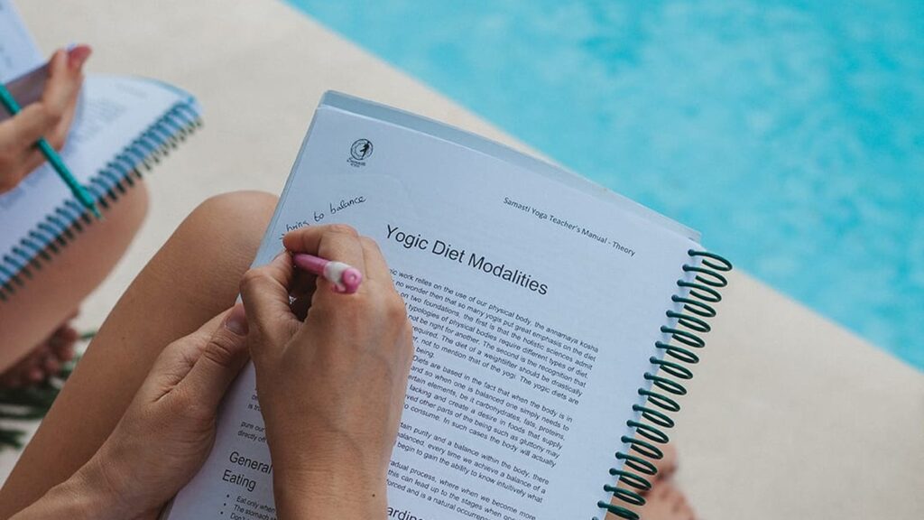A student studies the Samasti teacher training manual by the pool while researching a yoga teacher training in Bali